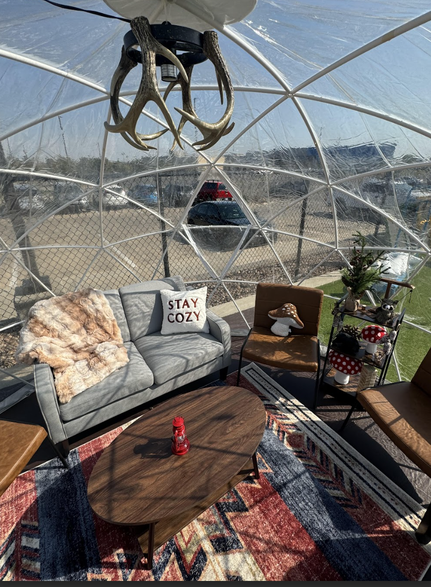 A cozy seating area inside a clear geodesic dome, featuring a gray sofa with a “Stay Cozy” pillow and a fur throw, a wooden coffee table with a red lantern, and rustic decor on a colorful patterned rug.